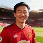 一位30岁左右男性球迷的半身肖像照片，他穿着曼联红色球衣，面带微笑，背景是比赛日的球场外景，阳光自然柔和。